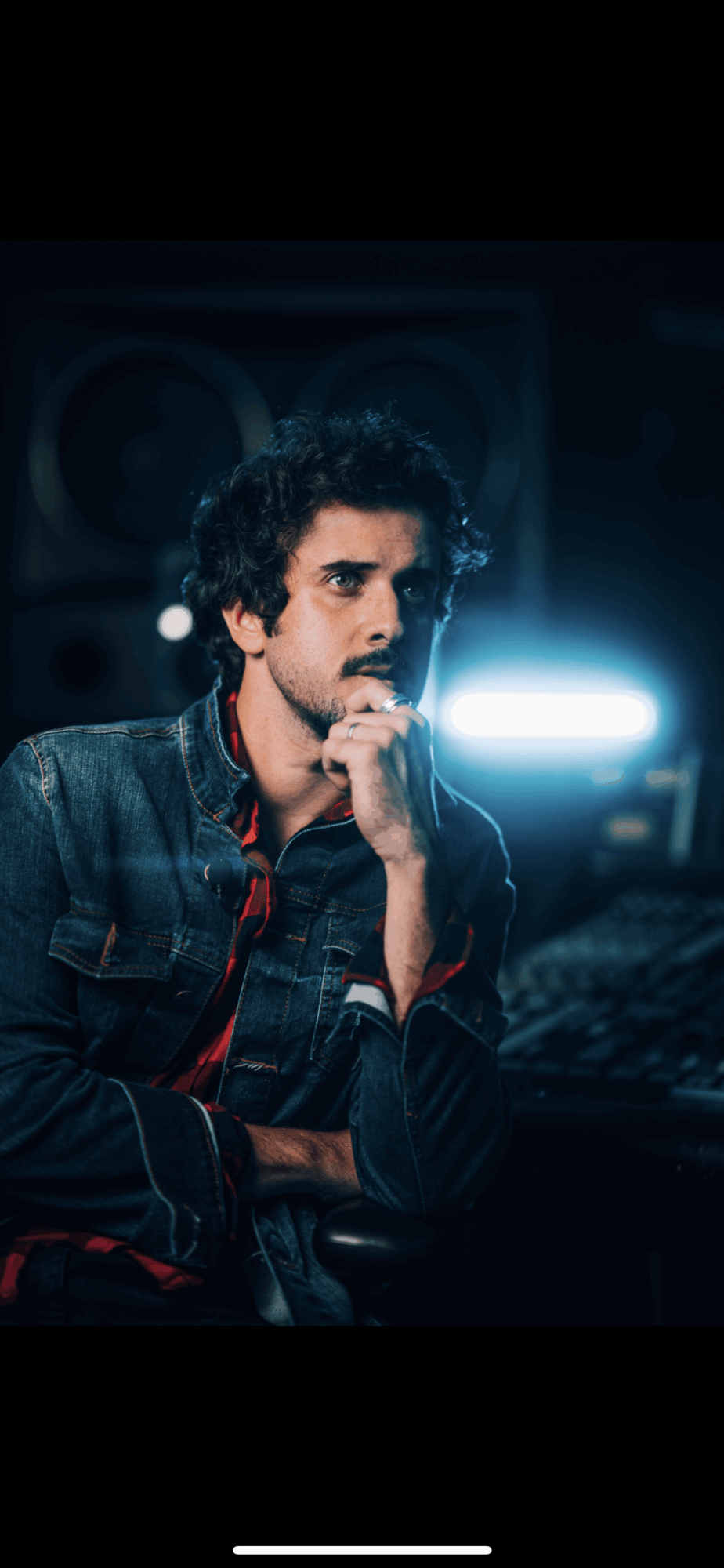Young man with curly hair and a beard, wearing a denim jacket, in a dark room with audio equipment, illuminated by a bright light.