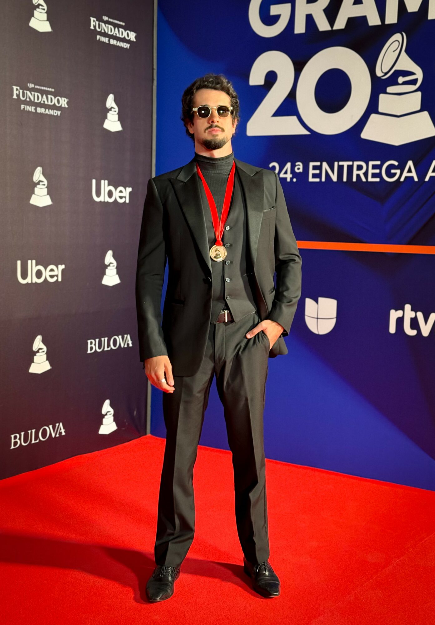 Man in a suit with sunglasses and a medal stands on a red carpet at an event backdrop.