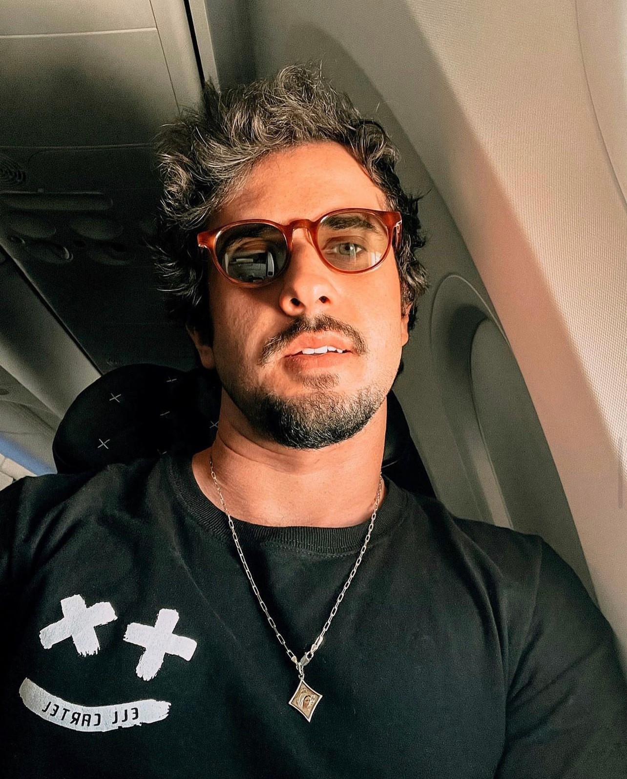 Man with curly hair, glasses, and a beard, sitting inside an airplane, wearing a black shirt and a necklace.
