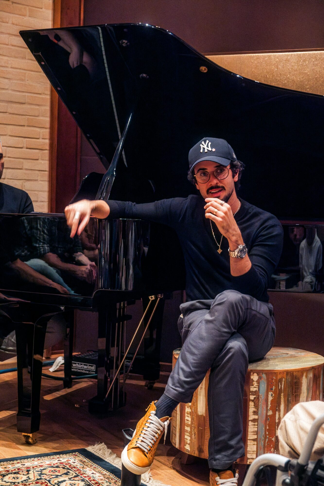 Person sitting on a stool, wearing a cap, sunglasses, and sneakers, holding a microphone near a grand piano in a room.