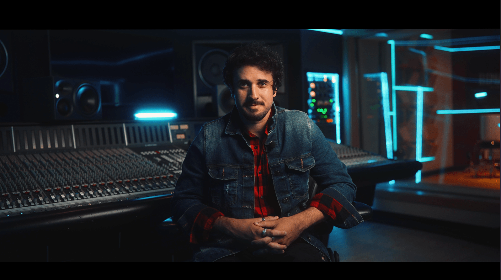 Man with curly hair and beard sitting in a music studio with equipment and blue lighting, wearing a denim jacket and red shirt.