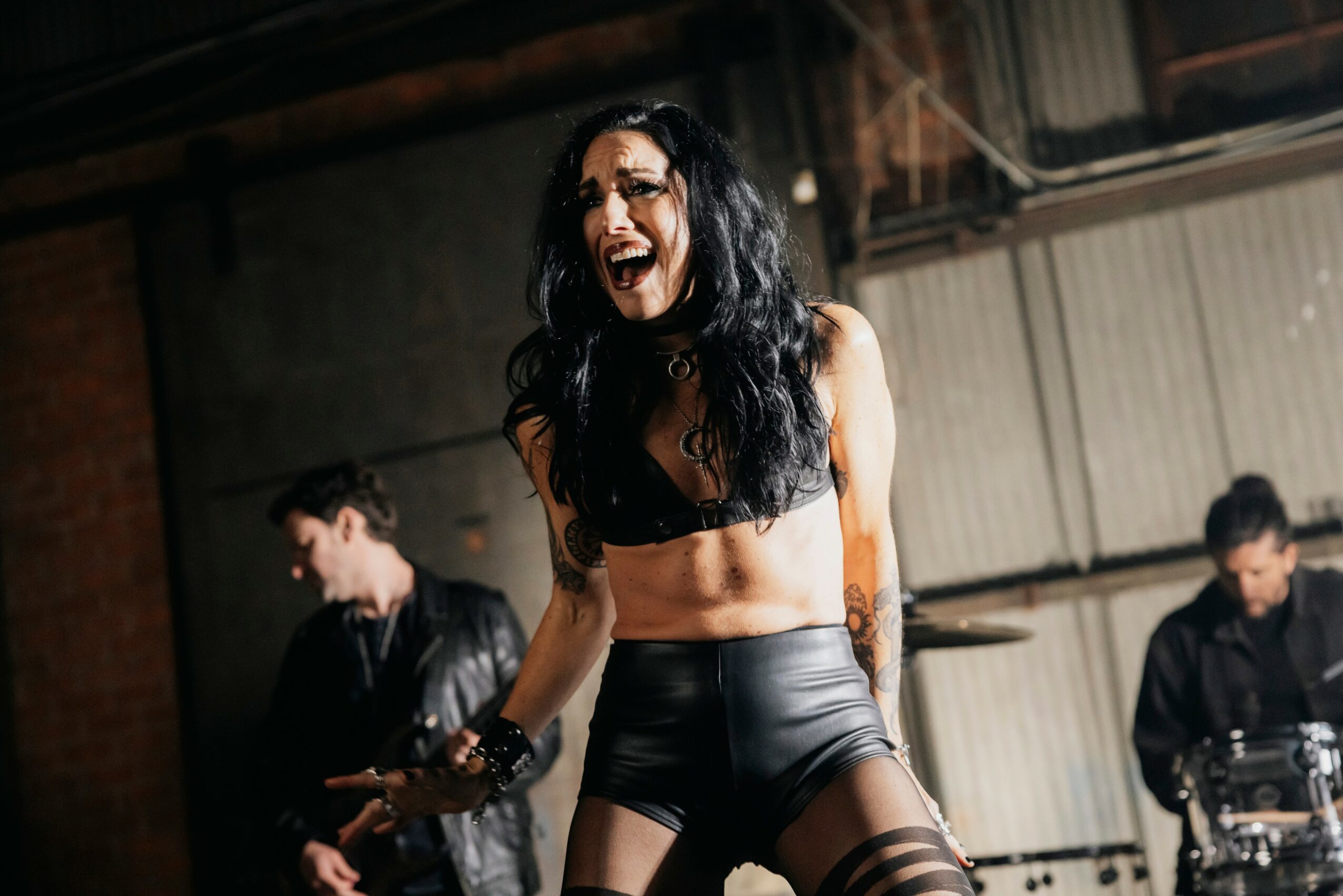 Woman with long dark hair laughing, wearing a black crop top and high-waisted shorts, in a dimly lit indoor setting.