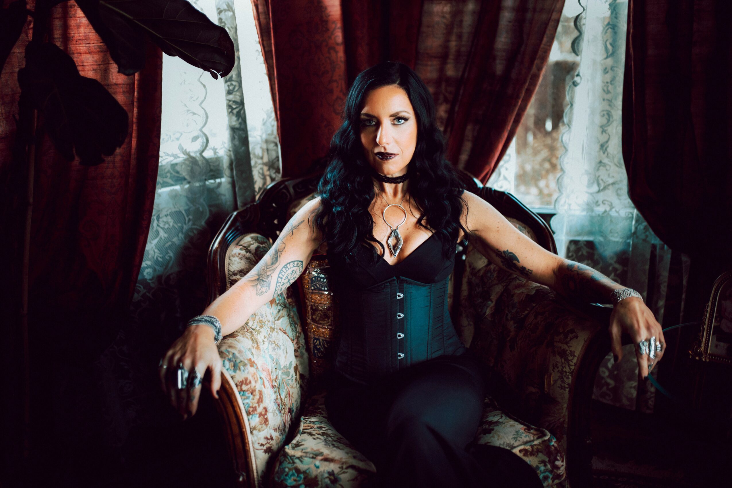 Woman with dark hair and makeup sitting on a vintage sofa in a dimly lit room with curtains.