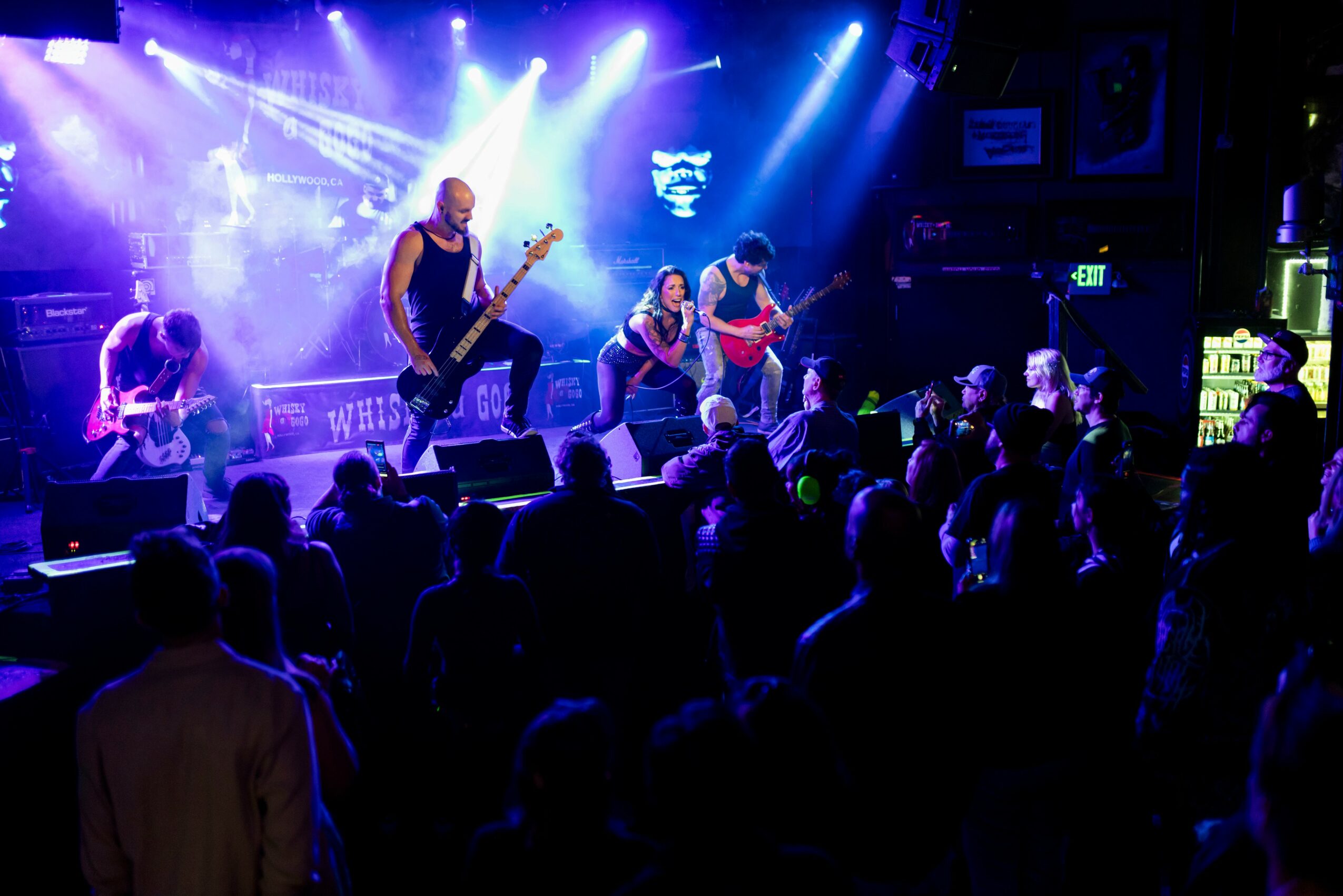 Band performs on stage with bright lights, audience watching in a dark venue, some silhouettes visible, lively atmosphere.