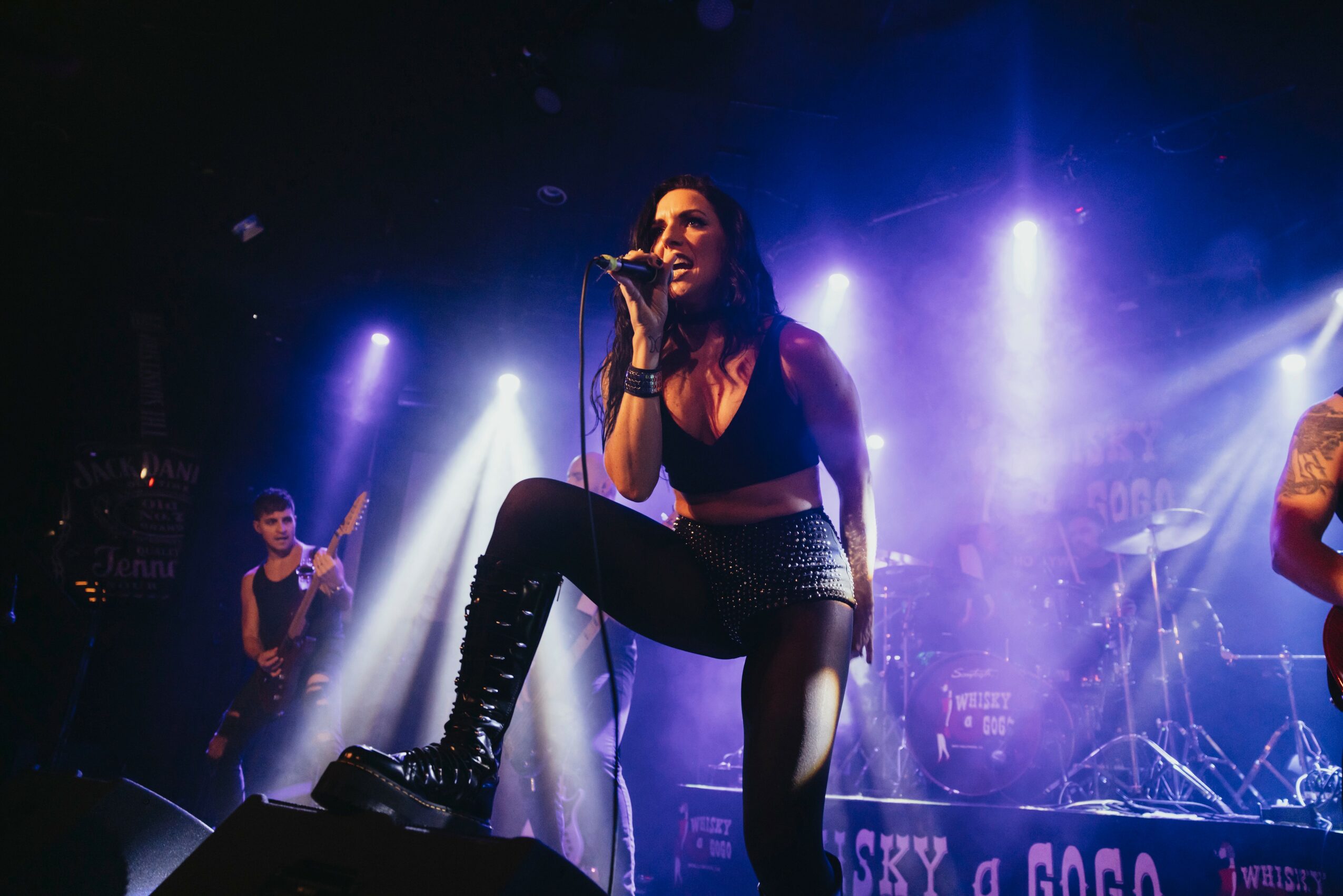 Female singer performing on stage with purple and blue lighting, holding a microphone, with band members in background.