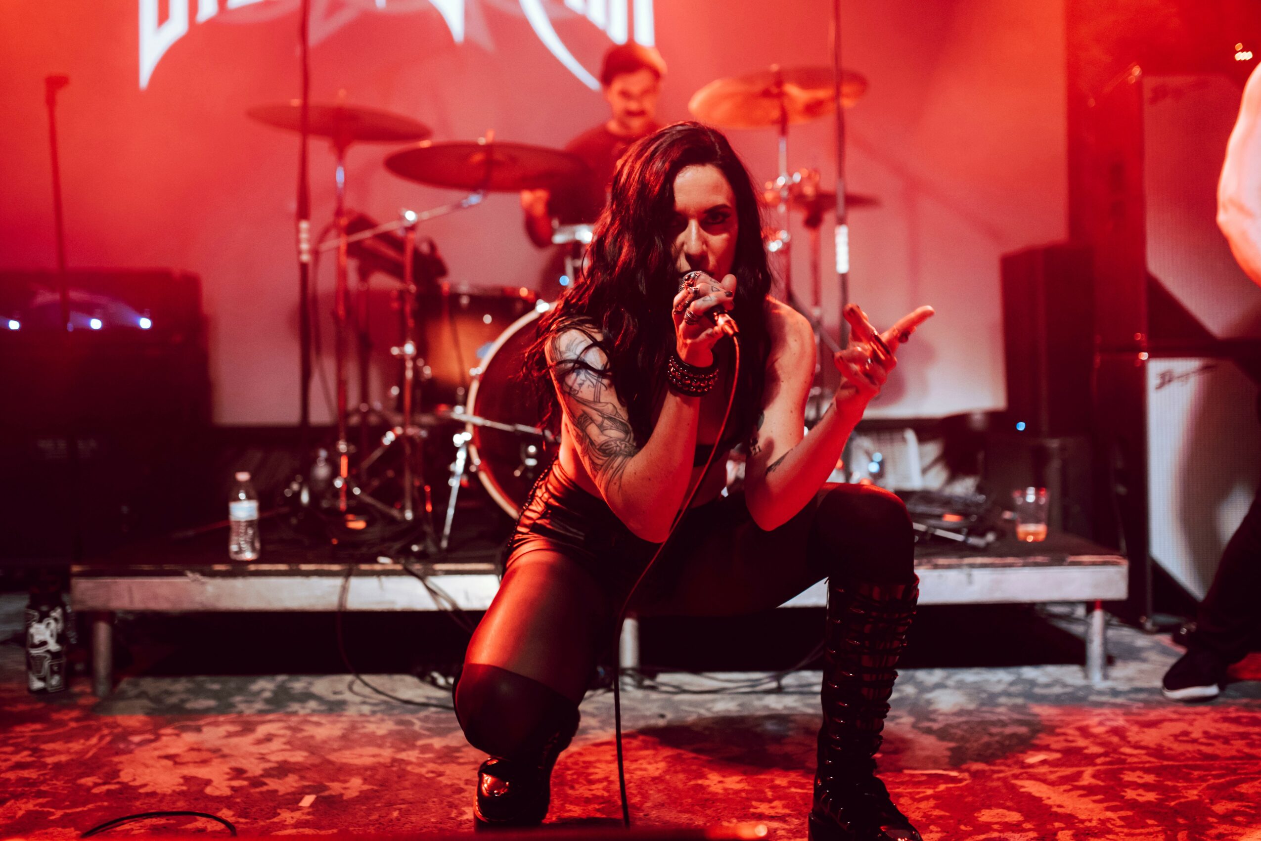 Female singer crouching on stage with a microphone, band playing in the background, red lighting, carpeted stage.