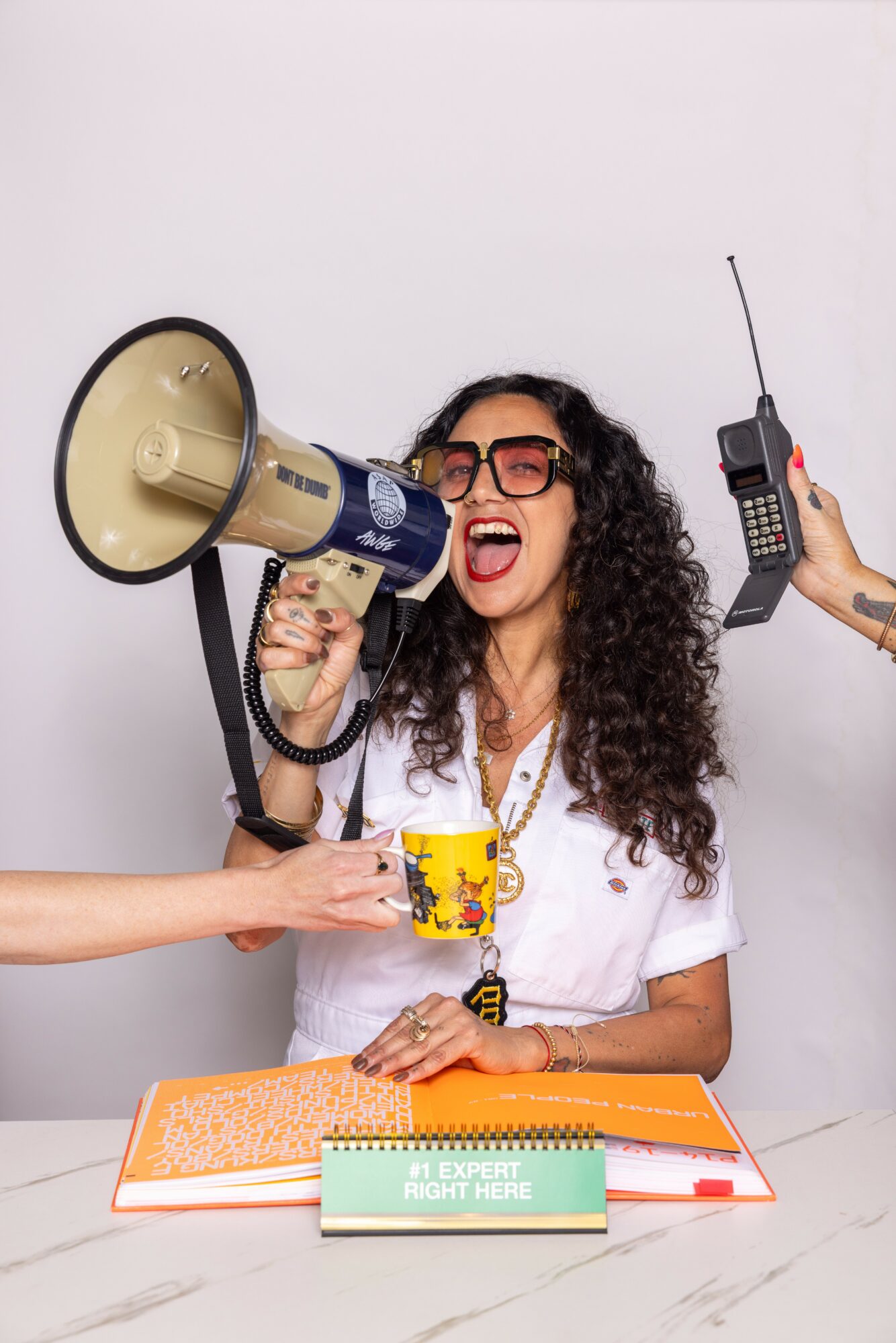 Woman with curly hair and glasses smiling, holding a megaphone, with people offering a mug and a walkie-talkie, sitting at a table.