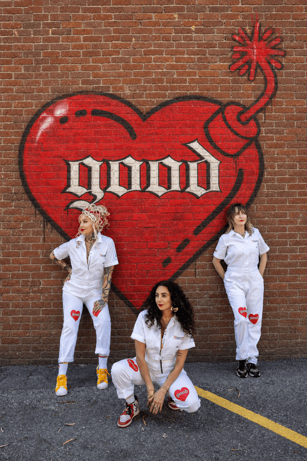 Three women in white outfits with red heart patches pose in front of a large red heart mural with a bomb and the word 'good'.