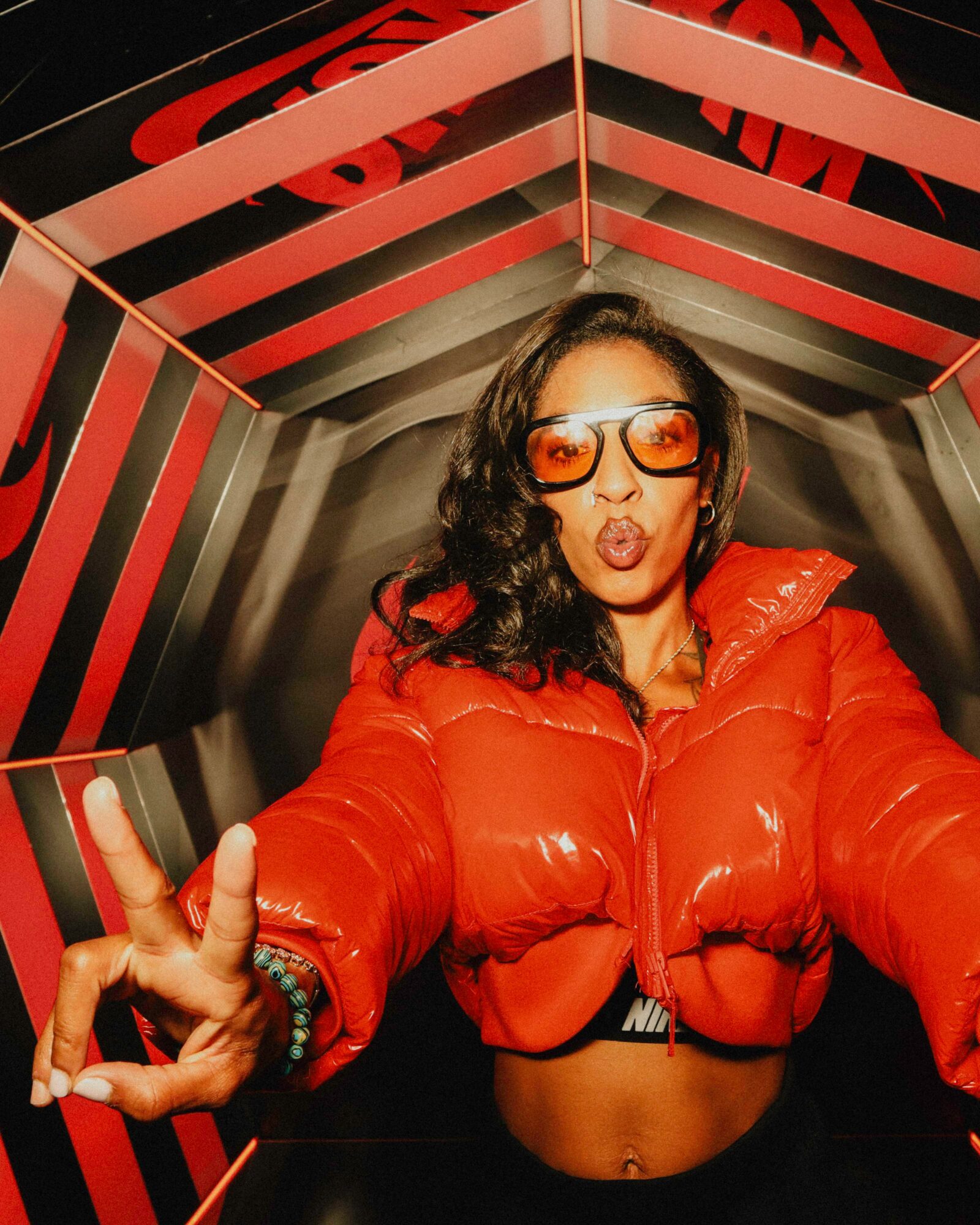 Woman with curly hair wearing sunglasses and a red jacket making a peace sign inside a geometric, colorful structure.