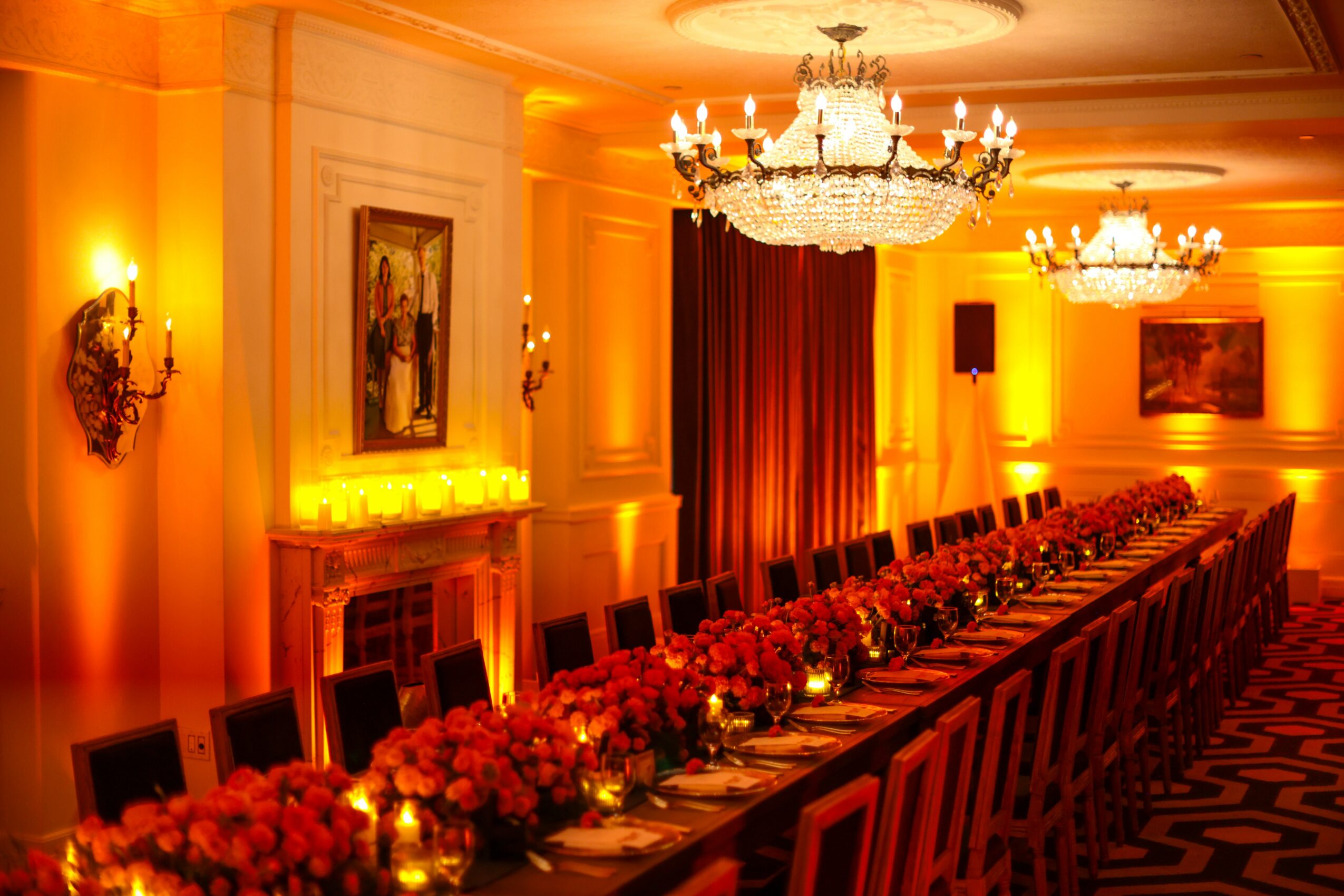 Long banquet table with floral centerpieces in a decorated room with chandeliers and warm lighting.