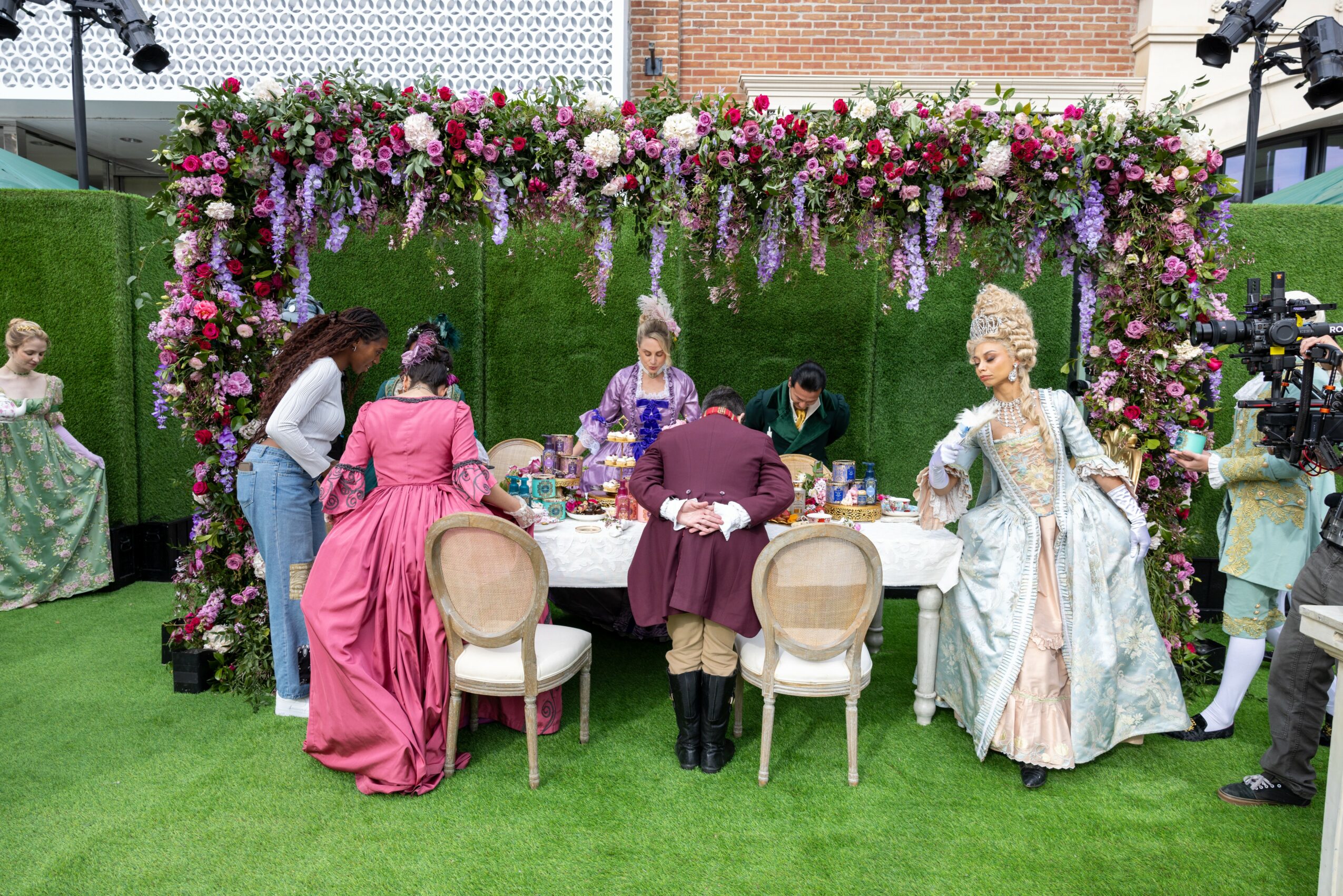 People in historical costumes gather around a table under a floral arch outdoors, with green grass and a hedge background.