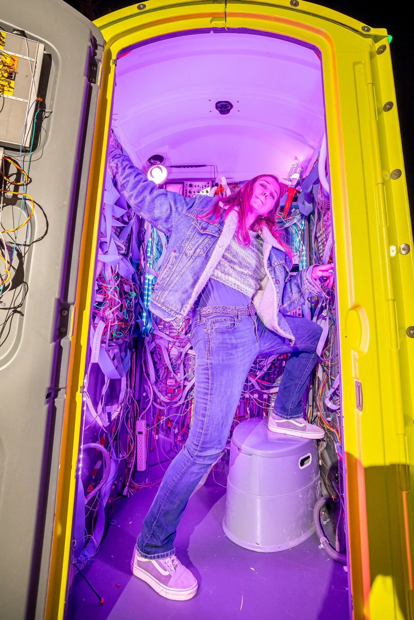Person with pink hair inside a colorful, illuminated capsule-like space with equipment and wires, holding onto the door frame.