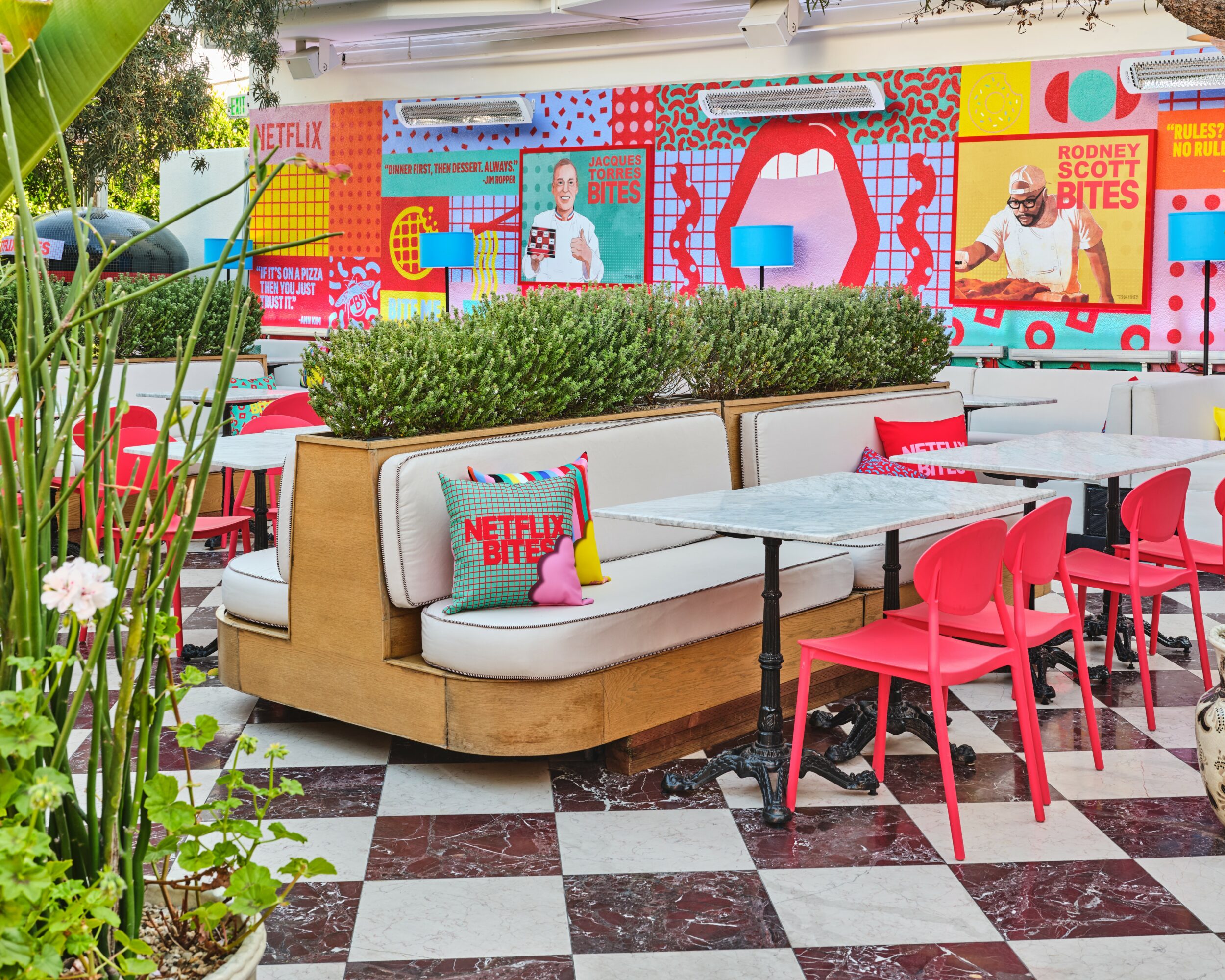 Indoor space with colorful wall art, a white cushioned bench, pink chairs, and checkered floor tiles.