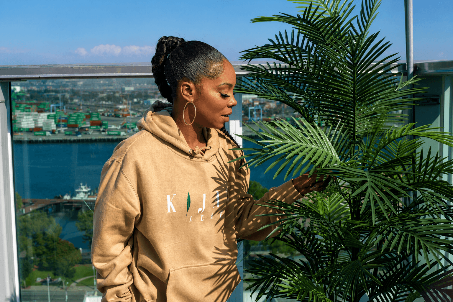 Woman with braided hair in a beige hoodie stands outdoors near a large plant, with a cityscape and water in the background.