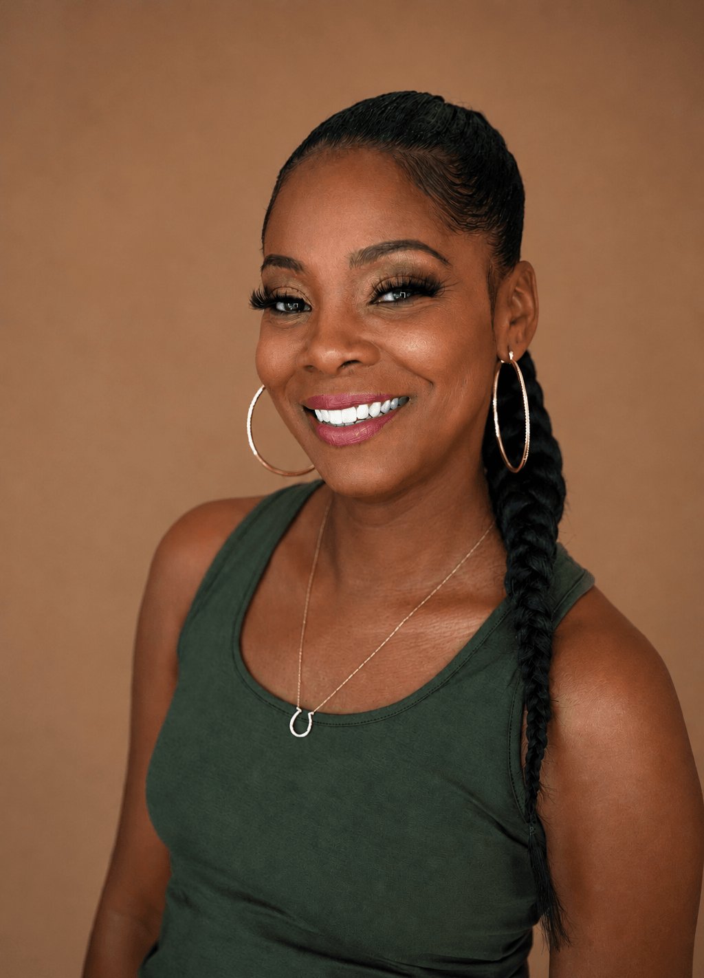 Smiling woman with braided hair, wearing hoop earrings and a green sleeveless top, against a plain background.