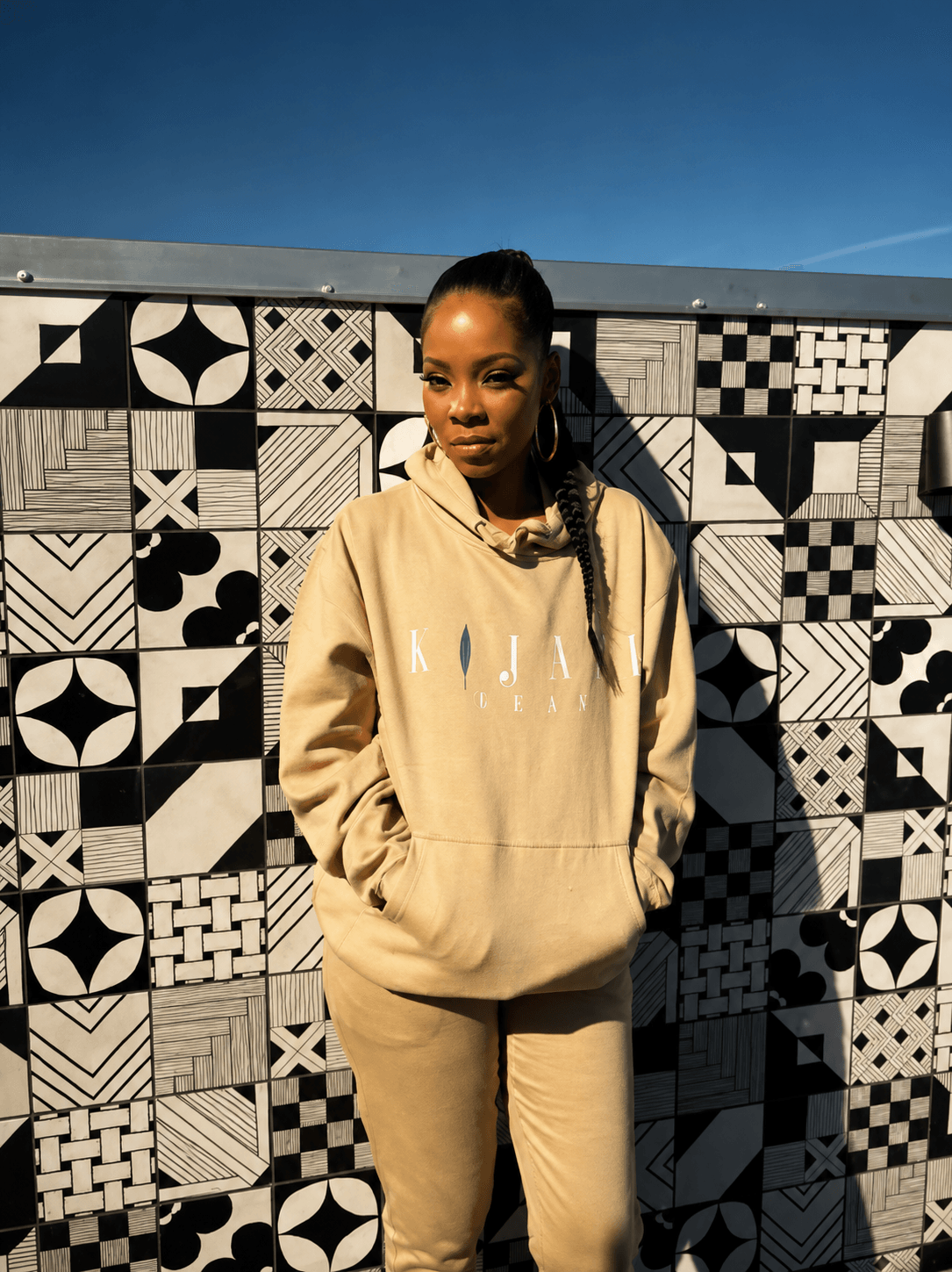 Young woman in beige hoodie and pants standing outdoors against a patterned wall, sunlight on her face.