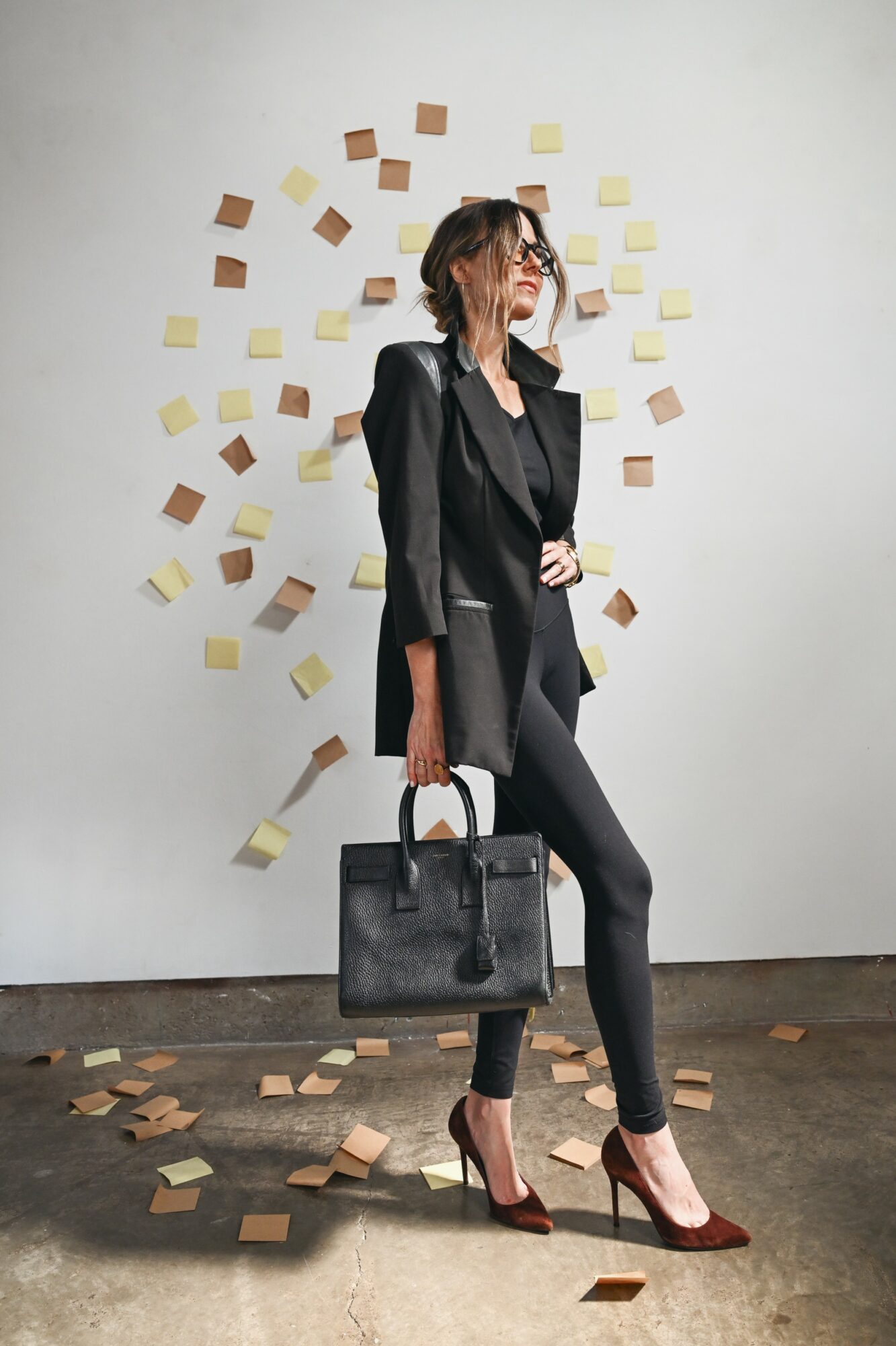 Woman in black blazer and pants holding a black handbag, standing against a wall with scattered sticky notes.