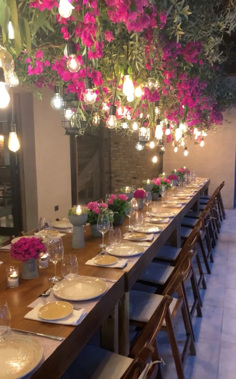 Long dining table decorated with pink and white flowers, set with plates, glasses, and napkins, under hanging pink flowers and lights.