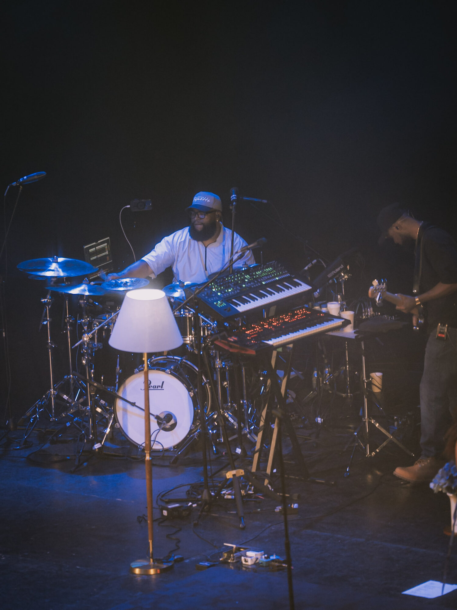 Musicians perform on stage with drums, keyboard, and guitar, illuminated by stage lights, dark background, and a floor lamp.