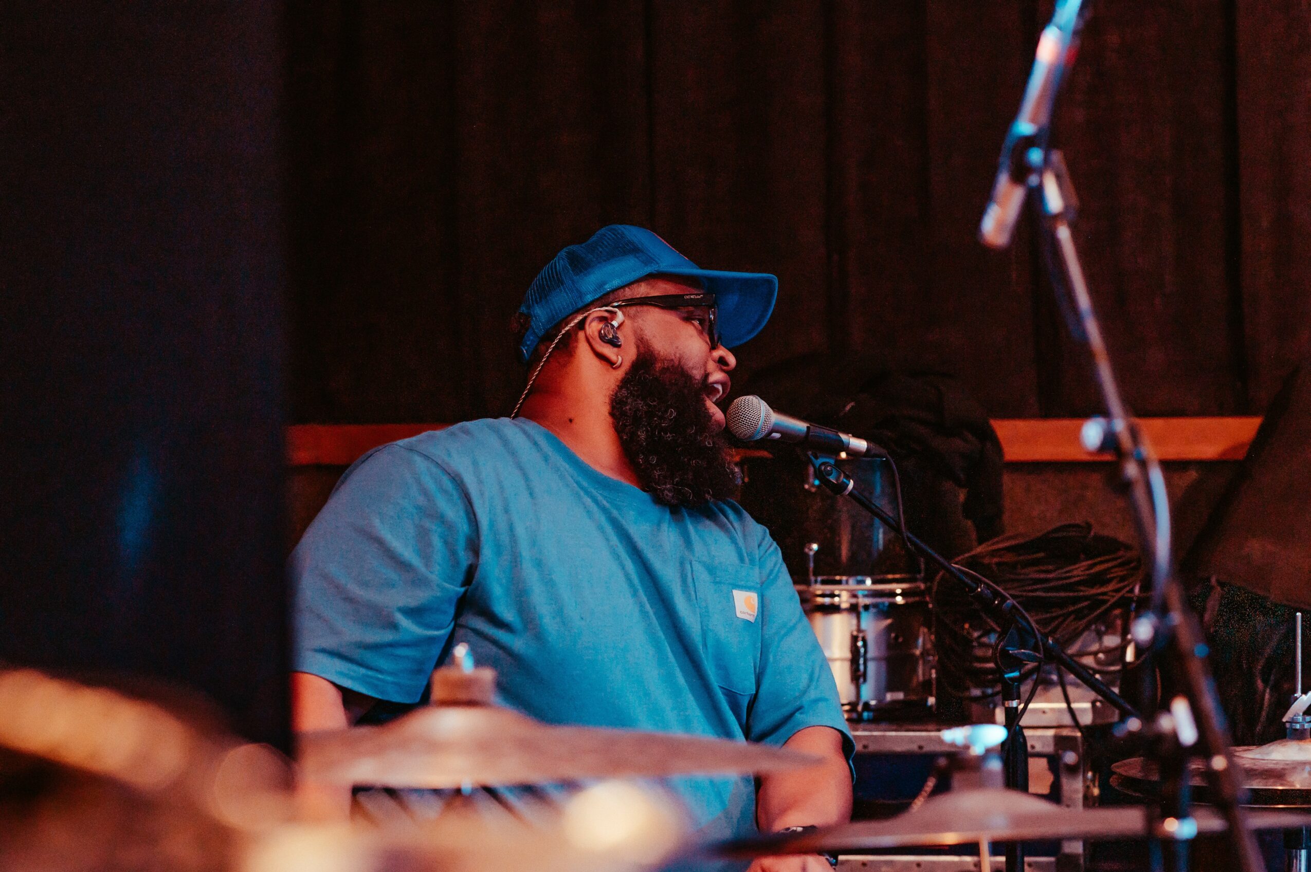 Bearded man in blue shirt and cap singing into microphone on stage with musical equipment, dark background.