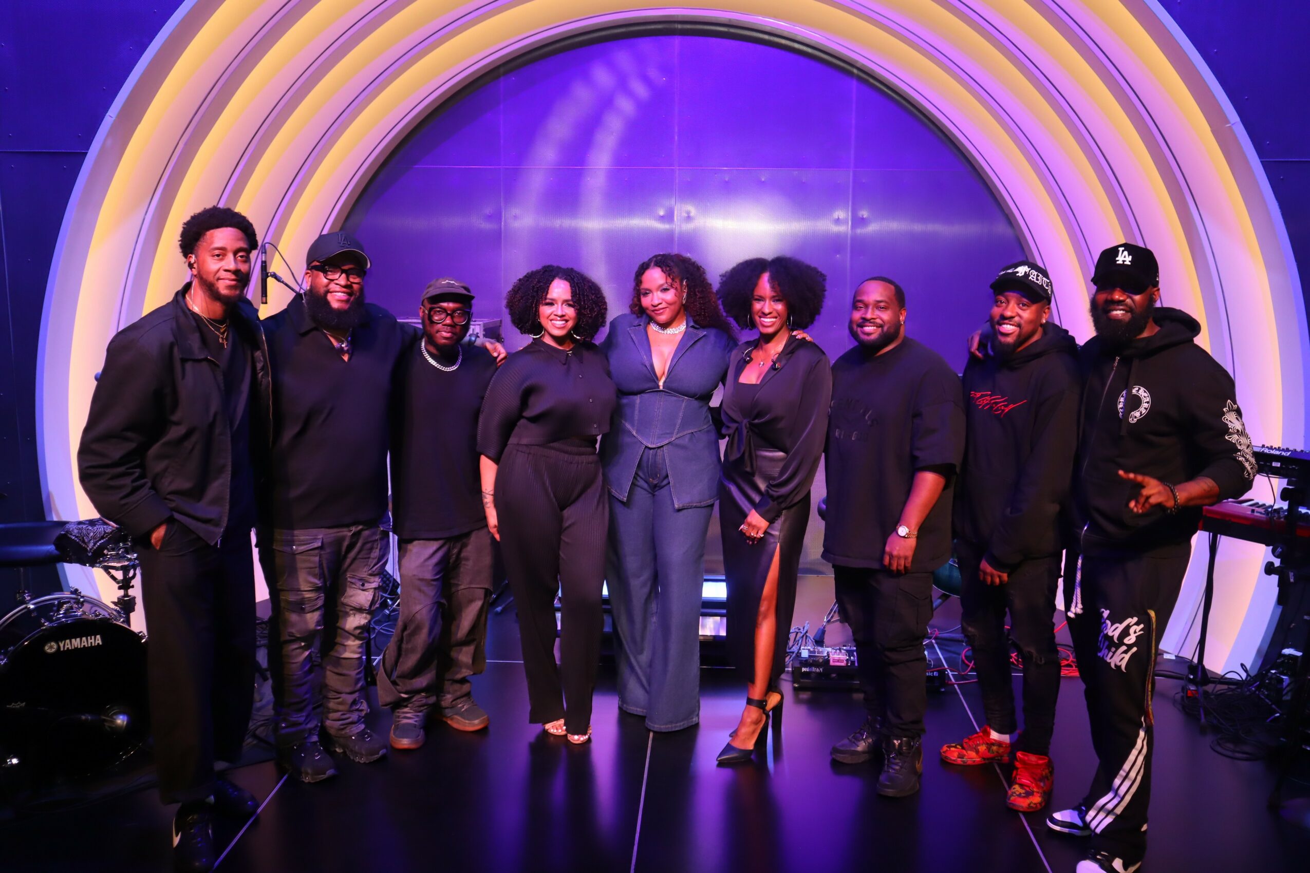 Group of nine people standing together in front of a colorful, circular background, smiling and posing for the photo.