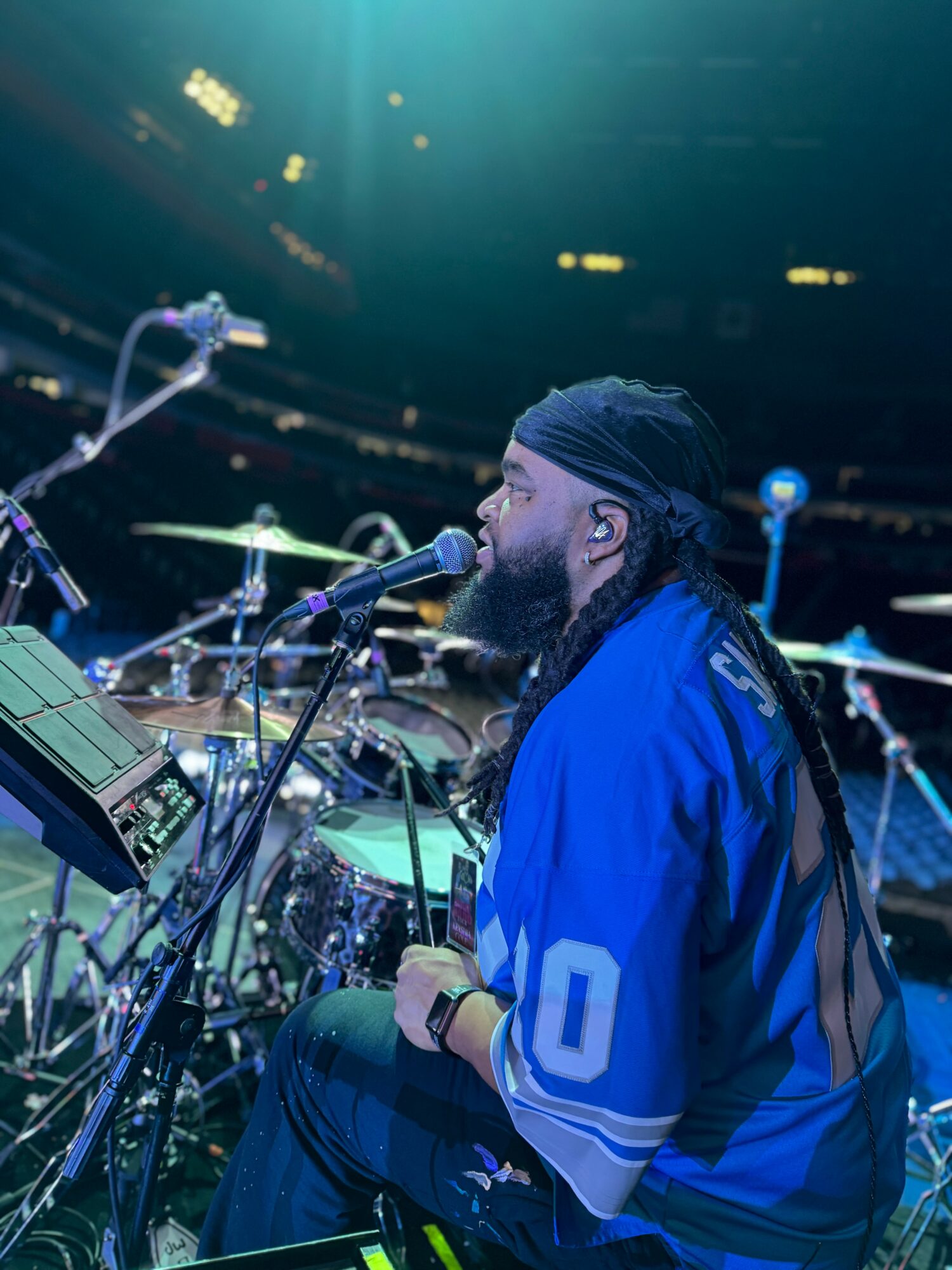 Musician with a beard and headband playing drums and singing into a microphone on stage.