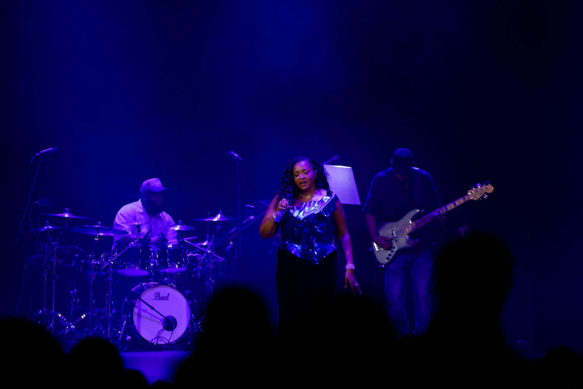 Three musicians perform on stage with blue lighting, one singing, one on drums, and one on guitar, audience visible.