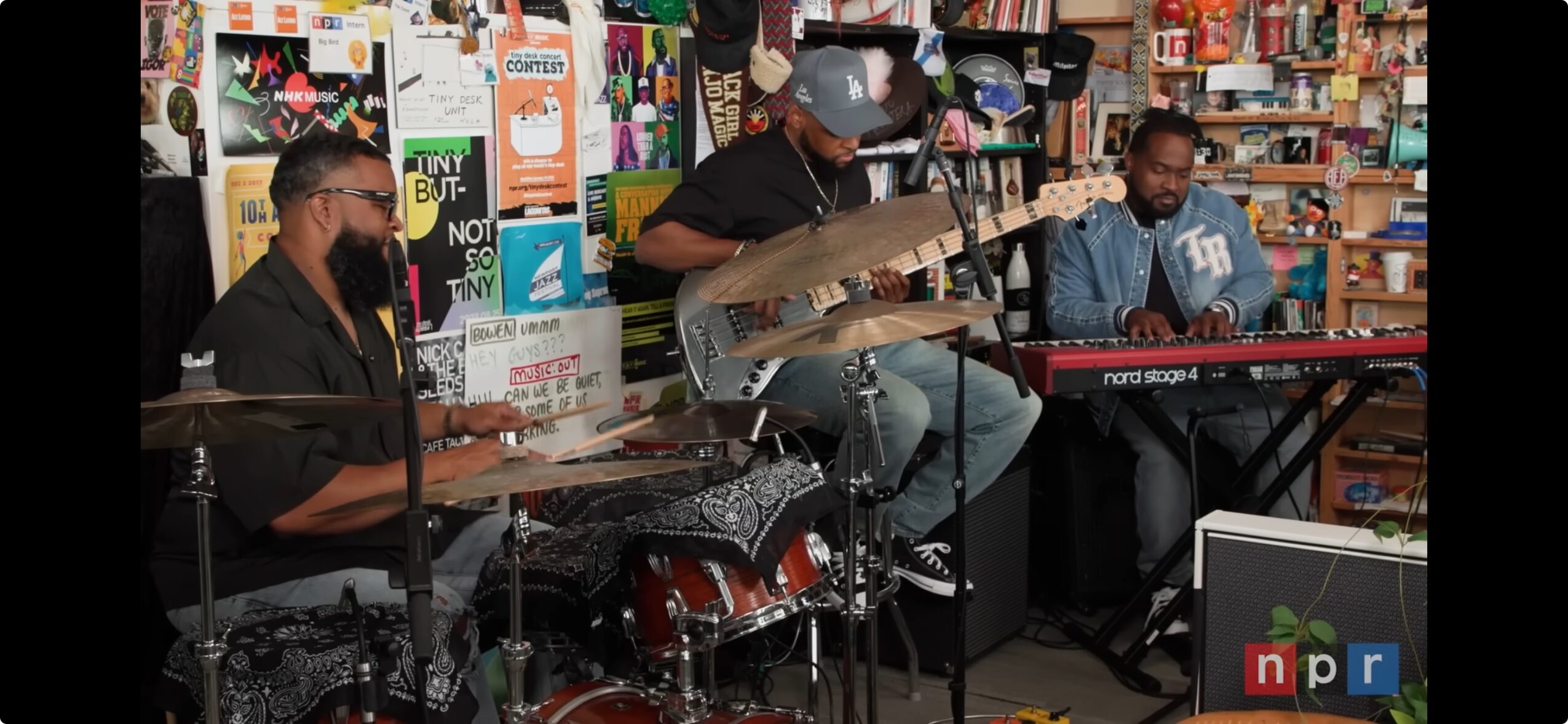 Three musicians perform with drums, guitar, and keyboard in a colorful store or cafe setting.