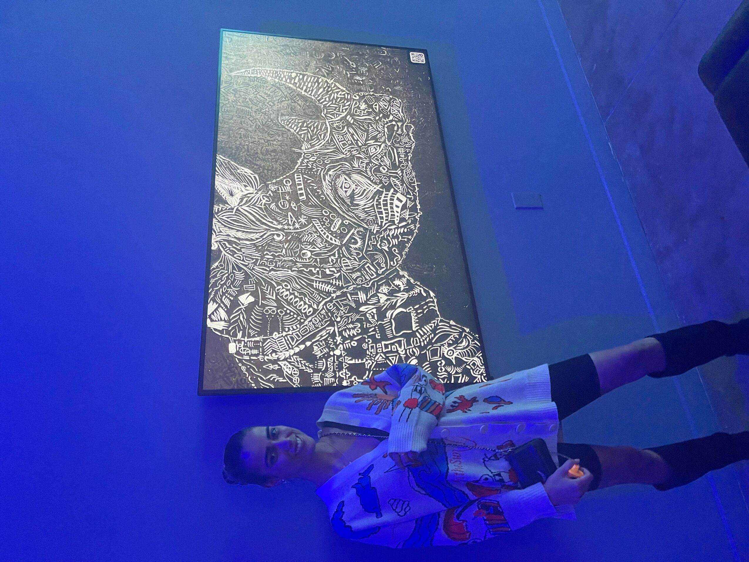 Person standing in front of a large, illuminated digital display with intricate white line art on a dark background, in a blue-lit room.