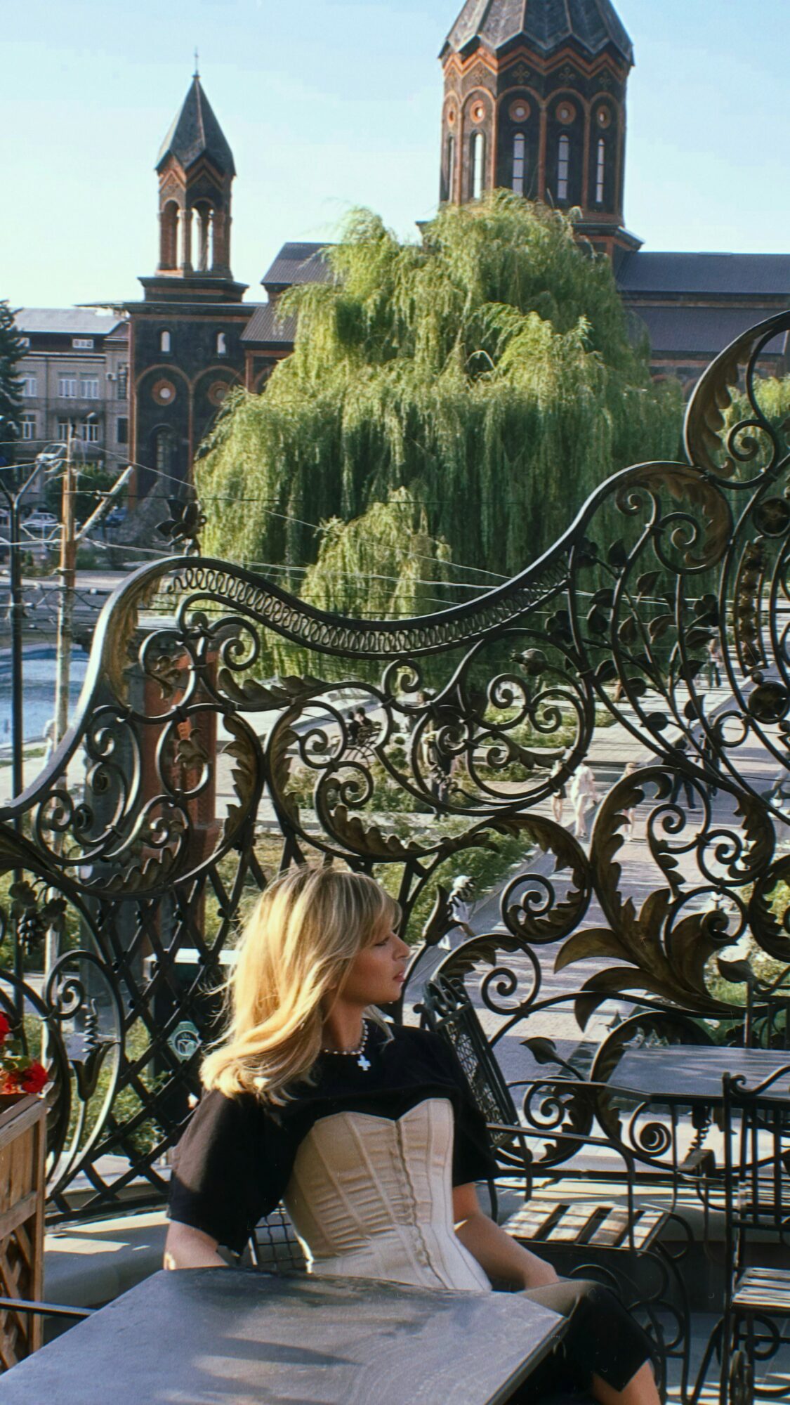 Woman with blonde hair sitting outdoors near ornate black metal railing, with a large tree and historic building in background.
