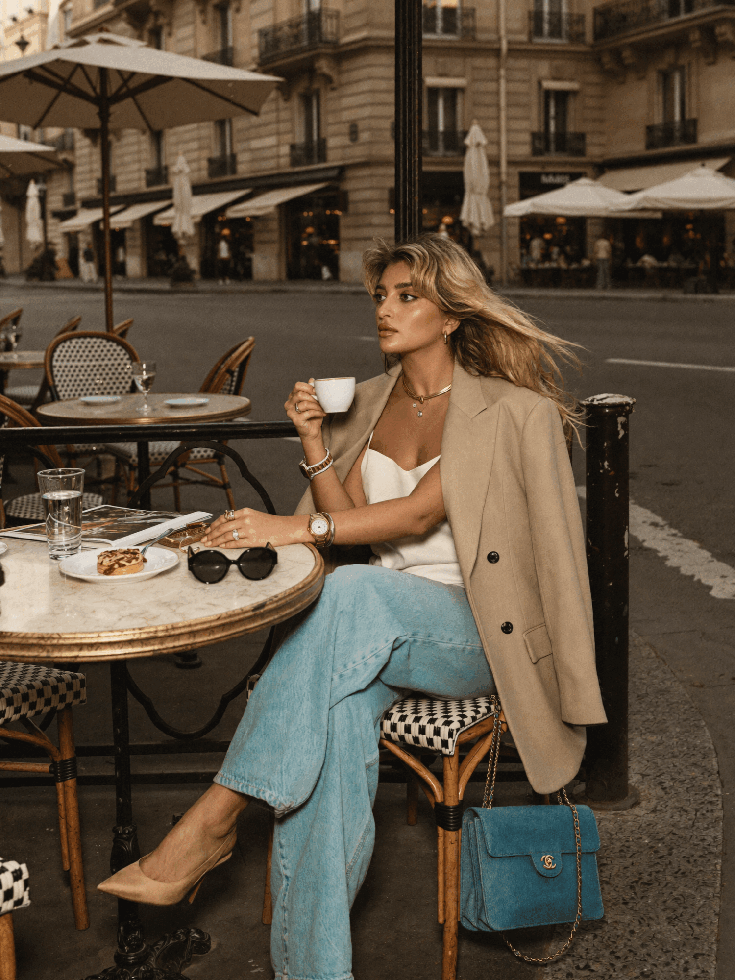 Woman sitting at an outdoor cafe, drinking from a cup, with a beige coat draped over her shoulders, on a city street.