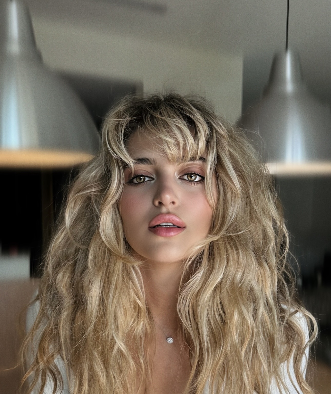 Young woman with long, wavy blonde hair and bangs, looking at the camera indoors, with pendant lights overhead.