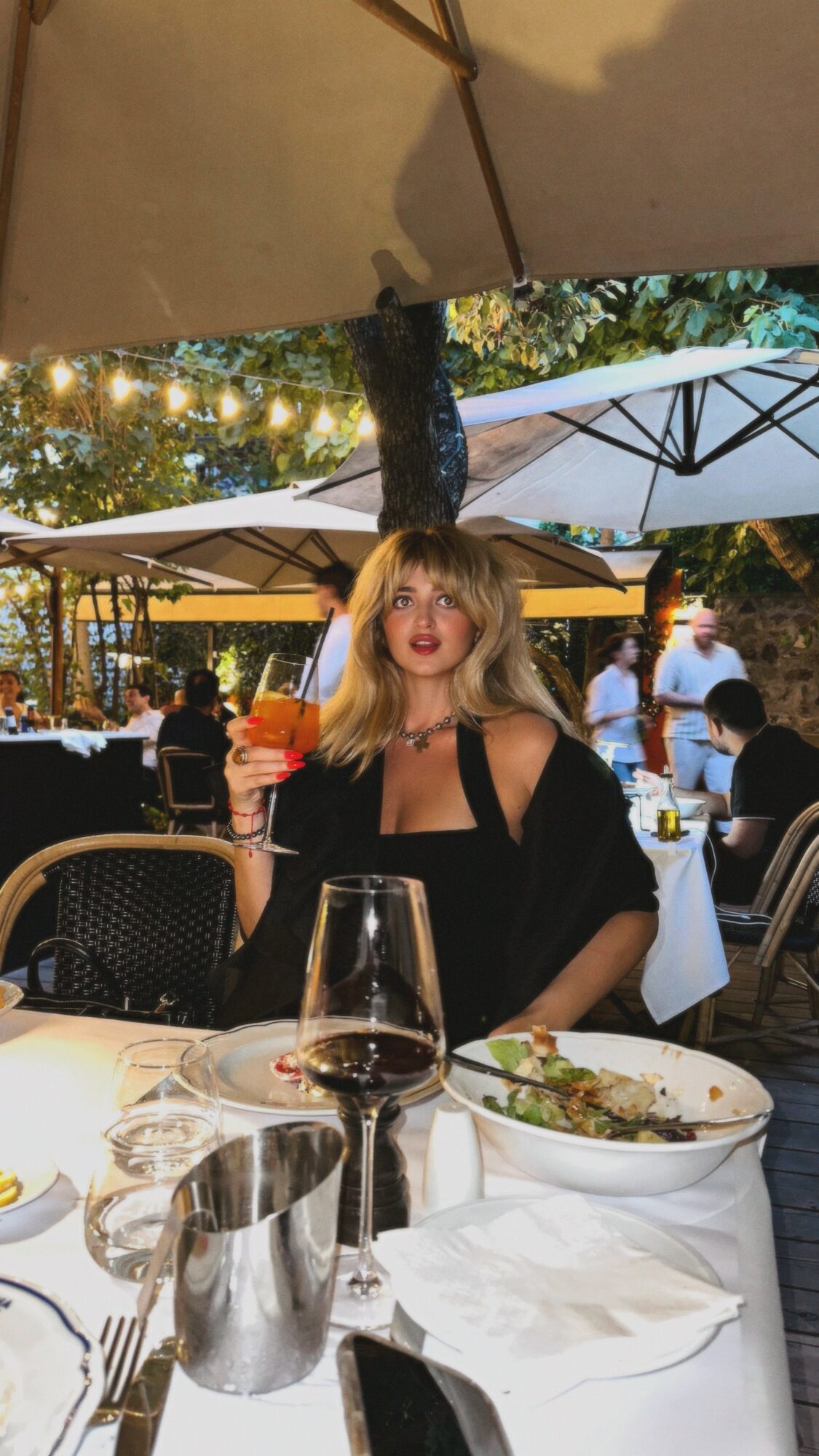 Woman with blonde hair holding a drink at an outdoor restaurant, surrounded by tables, umbrellas, and trees.