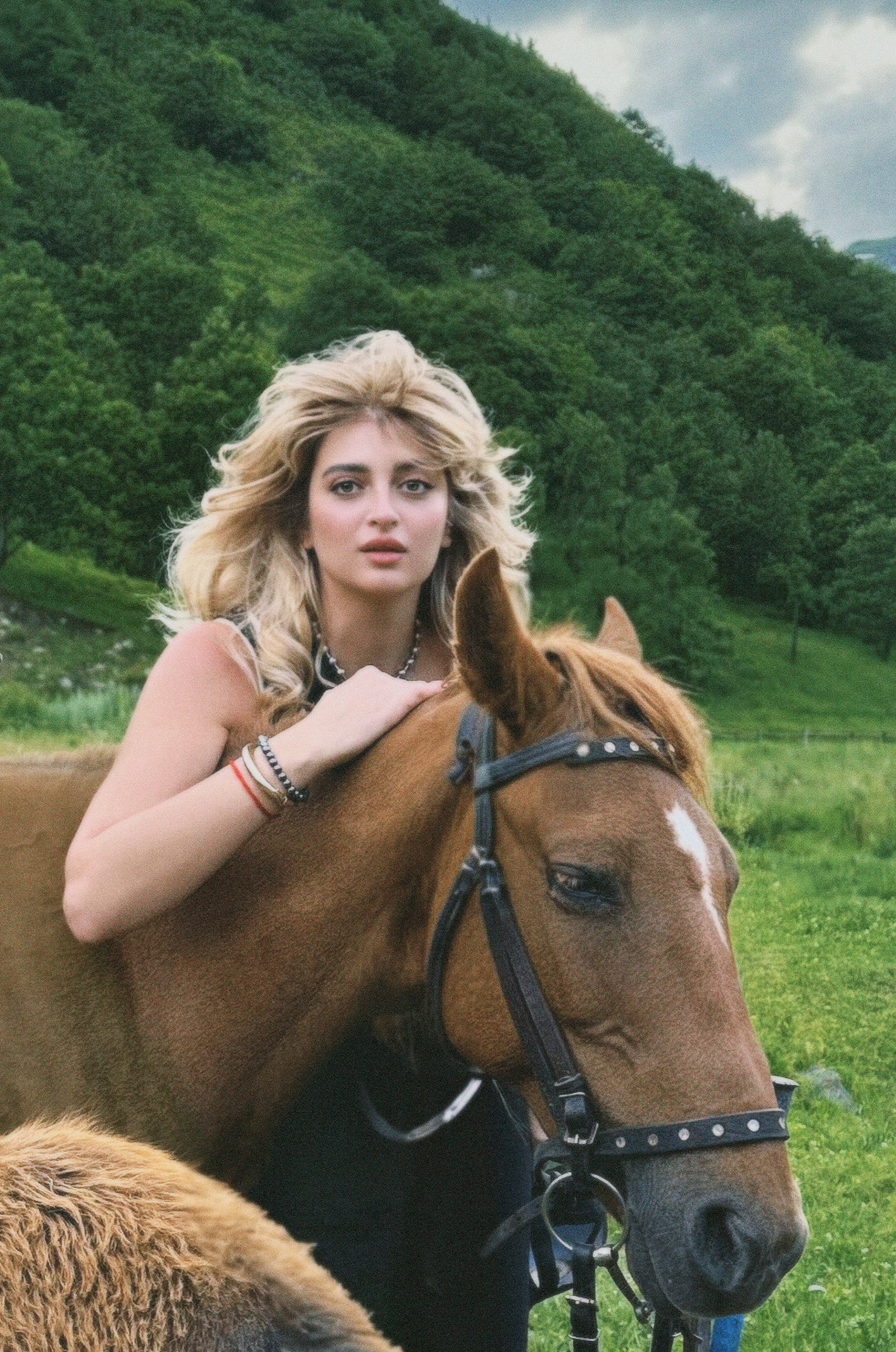 Young woman with blonde hair riding a brown horse outdoors with green hills and cloudy sky in background.