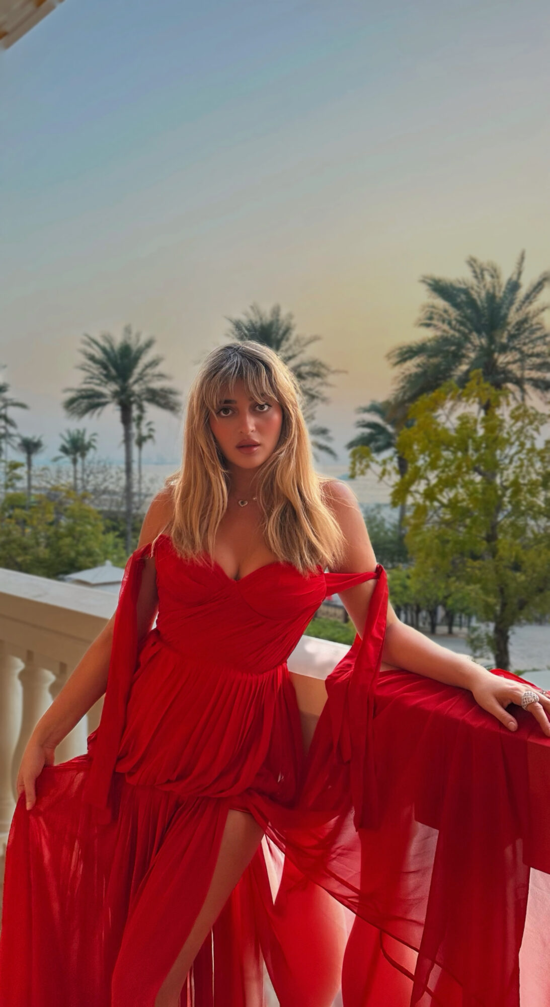 Woman in red dress standing outdoors with palm trees and a railing, sunlight in the background.