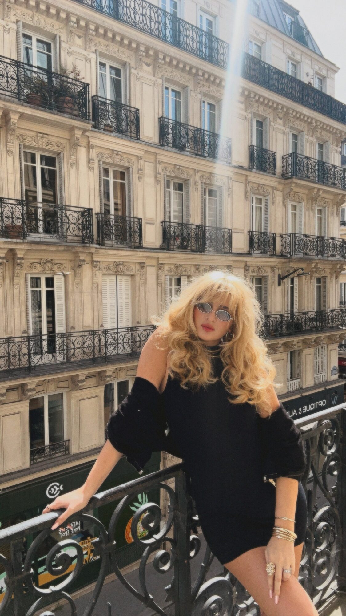 Woman with long blonde hair and sunglasses standing on a balcony with ornate black railing, Parisian building in background.