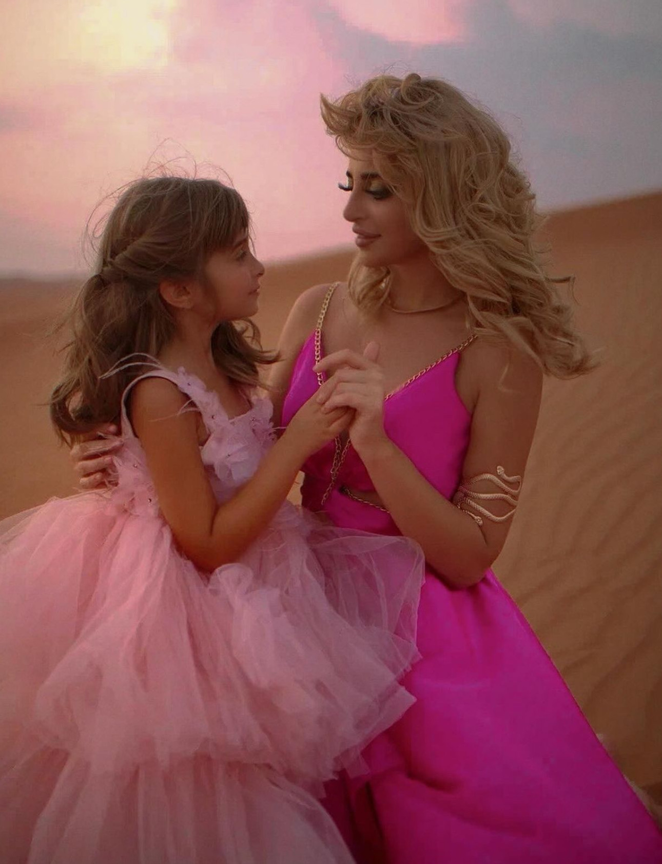 Woman and girl holding hands in a desert landscape during sunset, both wearing pink dresses, smiling at each other.