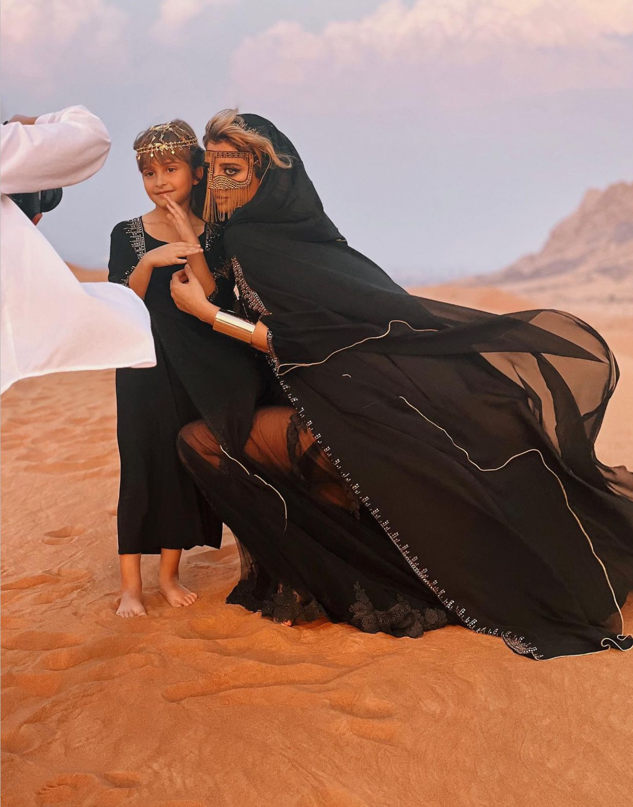Two women and a child standing on desert sand with mountains in background, woman in black traditional dress kneeling, child smiling.