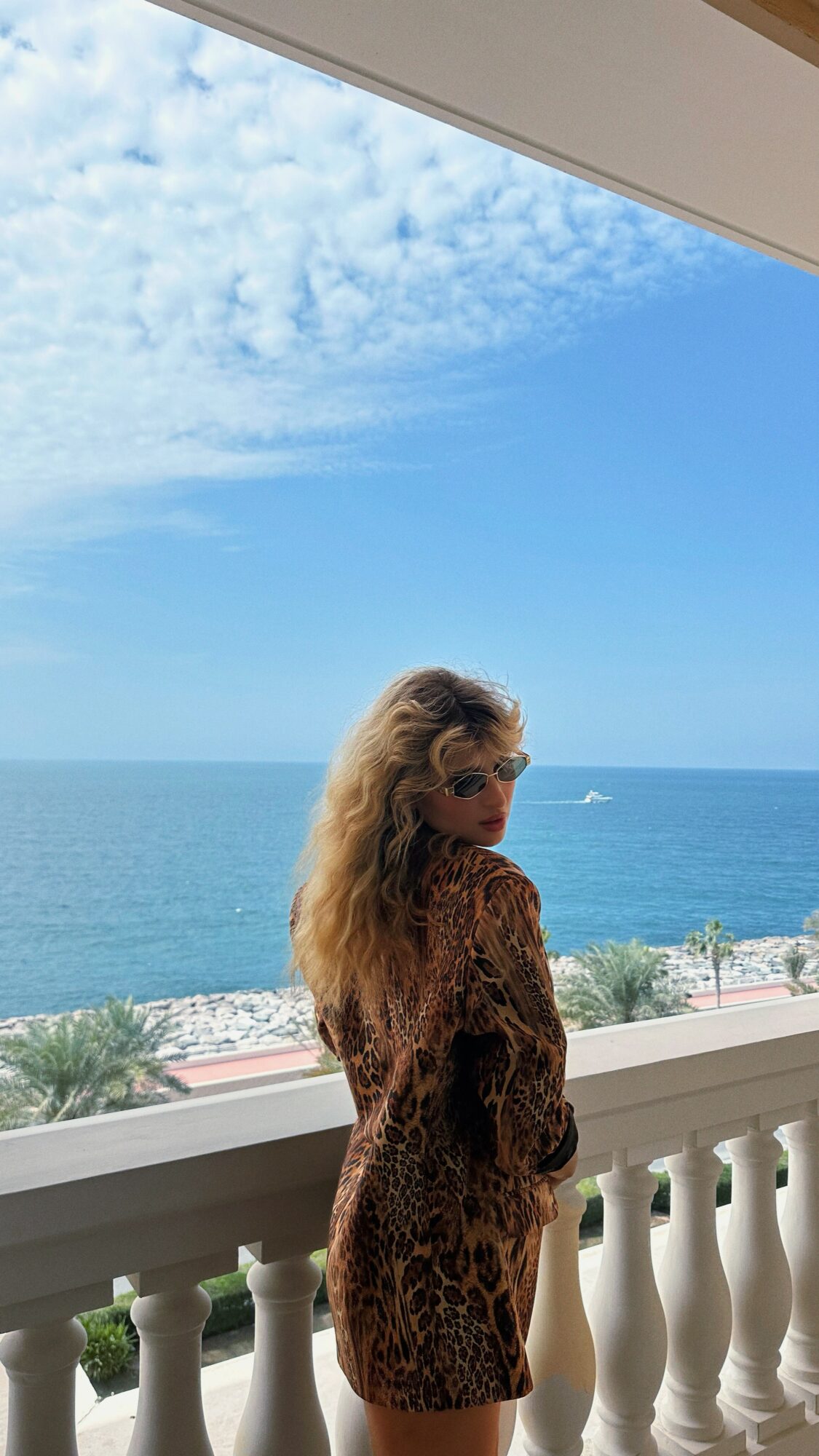 Woman with long hair standing on a balcony overlooking the ocean under a partly cloudy sky.