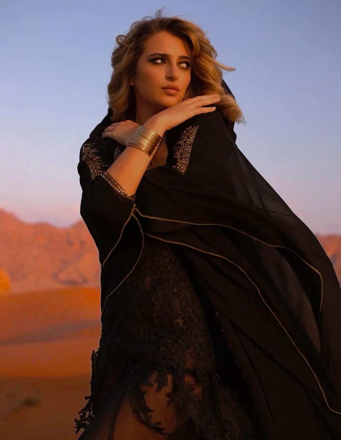Woman with wavy hair wearing black lace dress and gold bracelet, outdoors with desert landscape background.