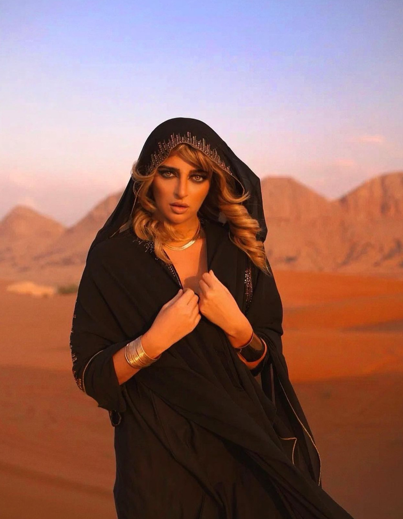 Woman in black hooded cloak standing in desert with mountains in background, sunset sky, holding the cloak near her chest.