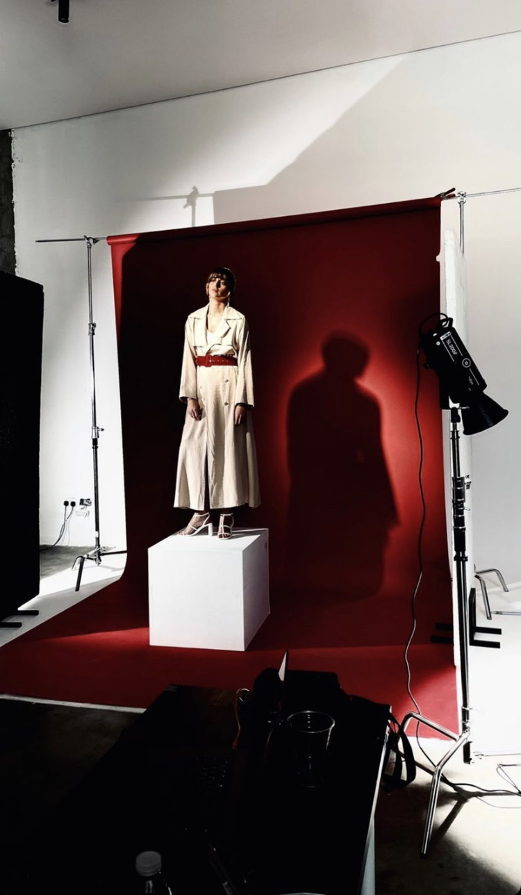 A mannequin dressed in a white outfit stands on a white pedestal against a red backdrop, with a shadow cast on the backdrop.