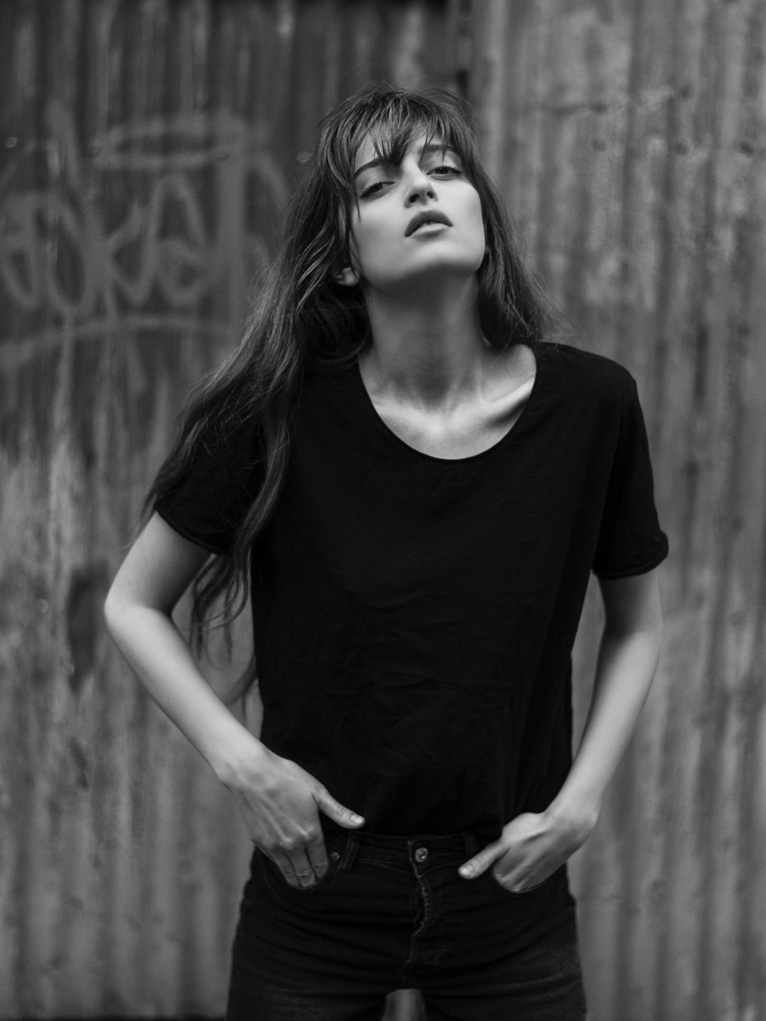 Young woman with long hair in a black t-shirt stands outdoors against a wooden fence, looking at the camera.
