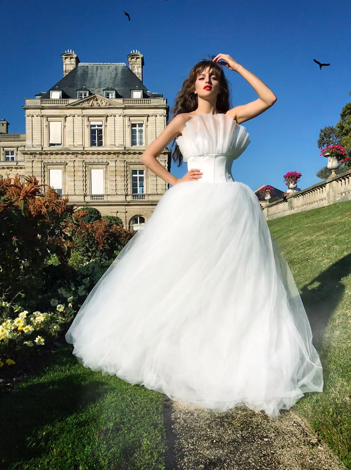 Woman in a white dress standing outdoors on grass with a historic building in the background, birds flying, and trees around.