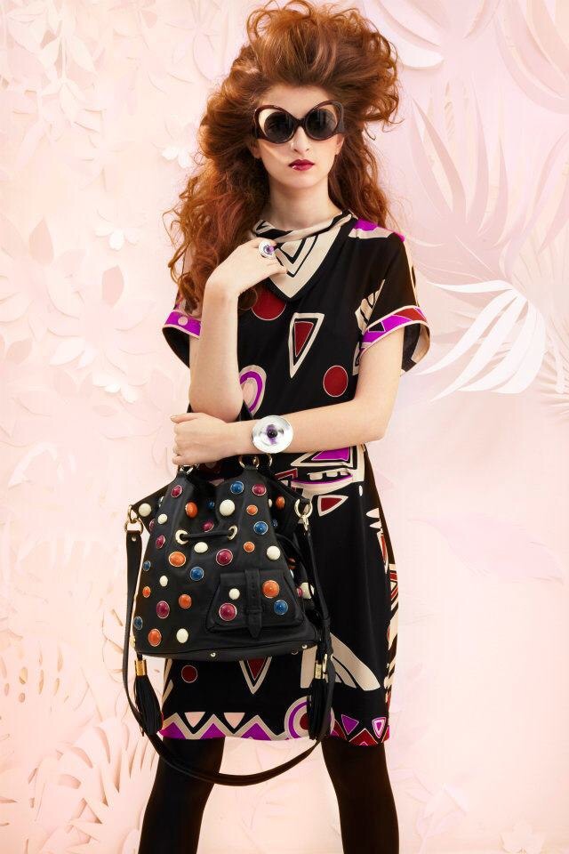 Woman with curly hair wearing sunglasses, a patterned dress, holding a polka dot bag, standing against a pink background.