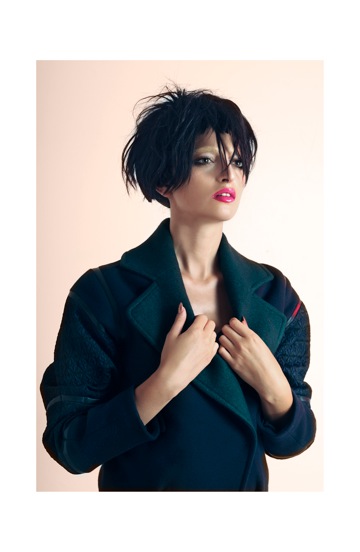 Woman with short black hair and red lipstick wearing a dark coat, holding the collar with both hands.