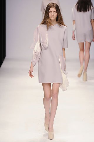 Model walking on runway wearing a light-colored dress and high heels, with other models in the background.