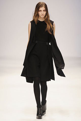 Young woman walking on runway wearing black sleeveless top, skirt, tights, and high-heeled shoes, holding a black coat.
