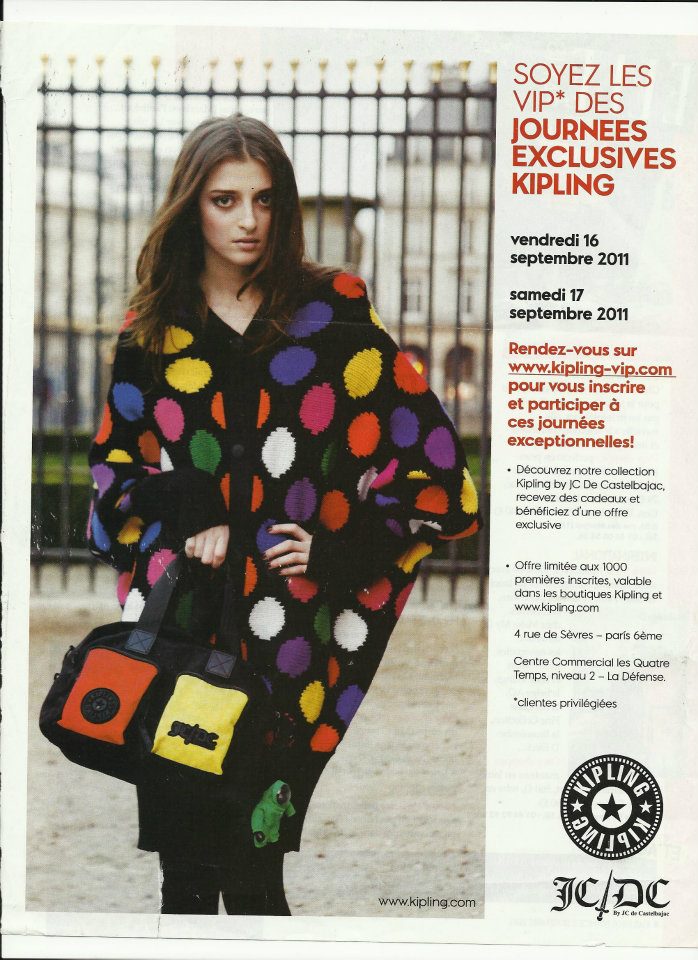 Young woman with long hair wearing a colorful polka dot coat, holding two bags, standing outdoors in front of a fence.