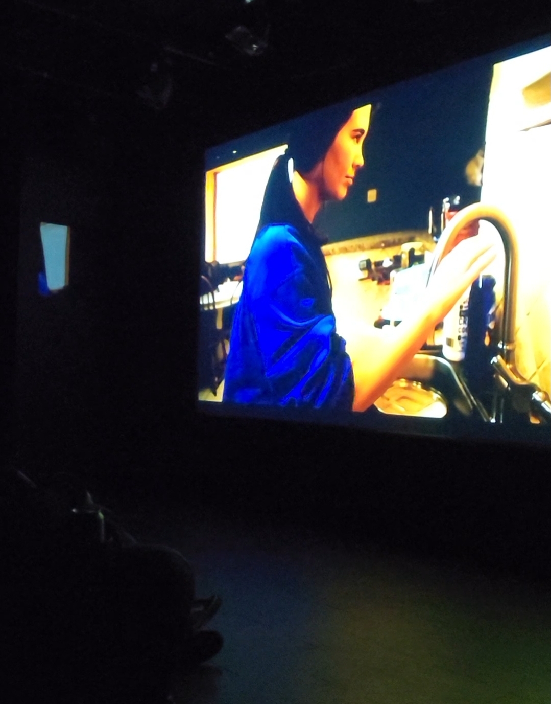 Person with dark hair in a blue shirt working on equipment, illuminated by a screen, in a dark room.